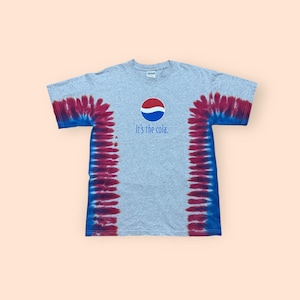 May include: A gray t-shirt with a tie-dye design in shades of red and blue on the sleeves and sides. The shirt features the Pepsi logo and the text "It's the cola." in blue.
