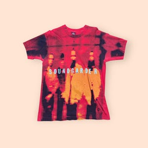 May include: Red Soundgarden t-shirt with a blurred image of the band members. The band's name is printed in light blue across the front. The shirt has short sleeves and a crew neck.