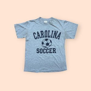 May include: A heather gray t-shirt with the words "CAROLINA SOCCER" printed in navy blue. A soccer ball graphic is centered between the words. The shirt is a crew neck style.
