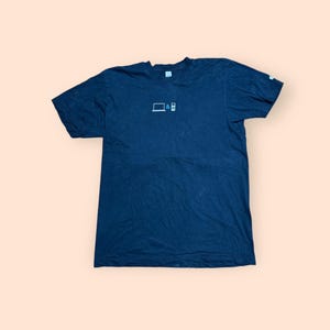May include: Dark navy blue t-shirt with a small white graphic of a laptop and a remote control. The shirt is made of a soft material and has short sleeves. The t-shirt is a casual wear item.
