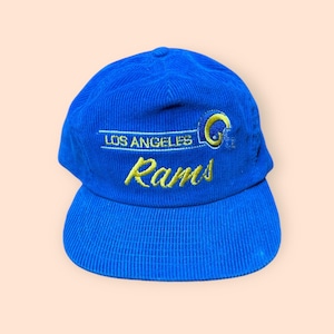 May include: A blue corduroy baseball cap featuring the text "LOS ANGELES" above the word "Rams" in yellow embroidery. A yellow football helmet graphic is on the right side of the cap.