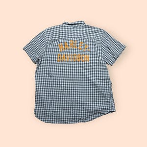 May include: A short-sleeved, button-down shirt with a green and white checkered pattern. The back of the shirt features the text "HARLEY DAVIDSON" in a yellow-orange color. The shirt has a classic collar and is made of a lightweight fabric.