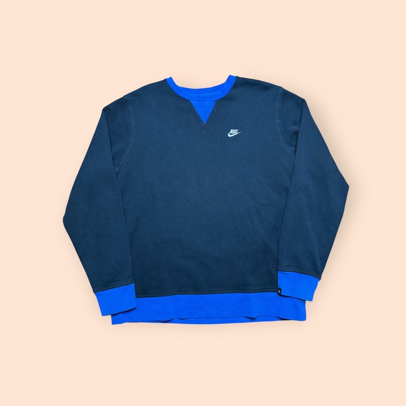 Puede incluir: Una sudadera azul marino con cuello y pu&ntilde;os azules. El logotipo de Nike est&aacute; bordado en blanco en la parte superior izquierda del pecho. La sudadera tiene cuello redondo y mangas largas.