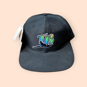 May include: Black baseball cap with a flat brim. The cap features an embroidered logo with the letters "TB" in a gradient of colors, with a stylized airplane below. A white tag is attached to the side.