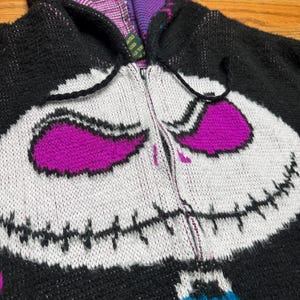 Puede incluir: Una sudadera de punto con cremallera en negro, blanco y morado que presenta un gr&aacute;fico grande de una cara de personaje con detalles de boca cosida. La capucha est&aacute; forrada con un patr&oacute;n morado y negro, y el frente incluye cremallera y cordones.