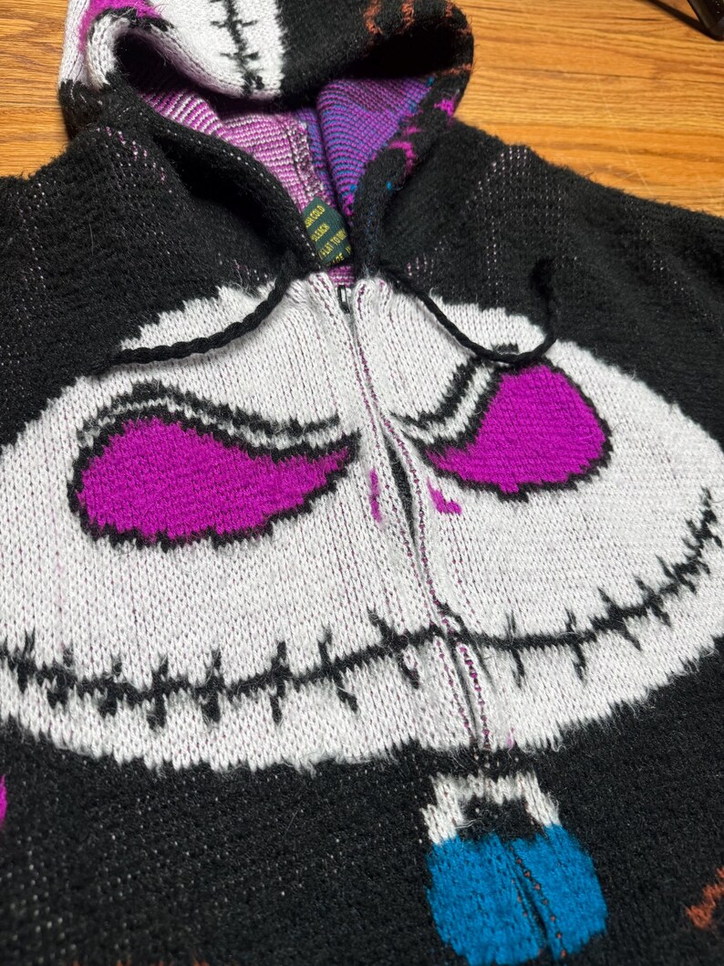 Puede incluir: Una sudadera de punto con cremallera en negro, blanco y morado que presenta un gr&aacute;fico grande de una cara de personaje con detalles de boca cosida. La capucha est&aacute; forrada con un patr&oacute;n morado y negro, y el frente incluye cremallera y cordones.