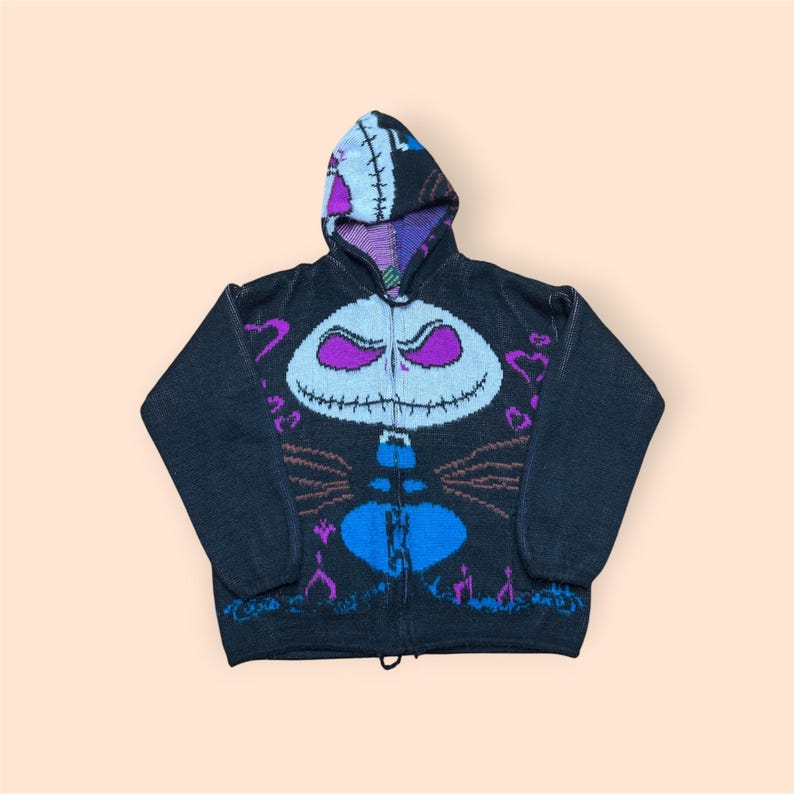 Puede incluir: Una sudadera de punto negra con cremallera y capucha que presenta un gr&aacute;fico grande de un personaje estilizado en la parte delantera. El dise&ntilde;o incluye una cara de calavera blanca con ojos morados, un torso azul y peque&ntilde;os corazones.