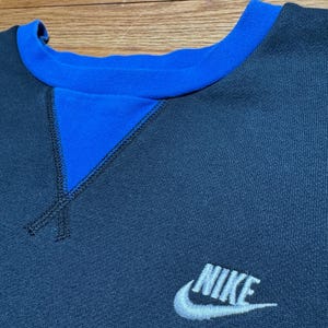 Puede incluir: Una sudadera azul marino con cuello azul y un detalle triangular azul en el escote. El logotipo de Nike est&aacute; bordado en blanco en la parte inferior derecha de la sudadera.
