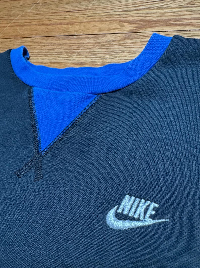 Puede incluir: Una sudadera azul marino con cuello azul y un detalle triangular azul en el escote. El logotipo de Nike est&aacute; bordado en blanco en la parte inferior derecha de la sudadera.