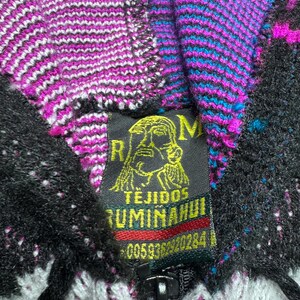 Puede incluir: Una sudadera de punto con cremallera y un patr&oacute;n vibrante en negro, blanco, magenta y turquesa. Una etiqueta de tela negra cosida en el cuello muestra un gr&aacute;fico amarillo de un rostro y el texto RM TEJIDOS RUMI&Ntilde;AHUI 0059362920284.