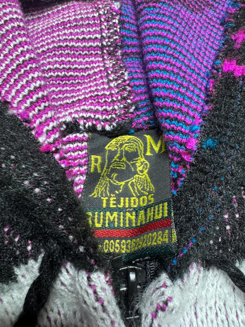 Puede incluir: Una sudadera de punto con cremallera y un patr&oacute;n vibrante en negro, blanco, magenta y turquesa. Una etiqueta de tela negra cosida en el cuello muestra un gr&aacute;fico amarillo de un rostro y el texto RM TEJIDOS RUMI&Ntilde;AHUI 0059362920284.