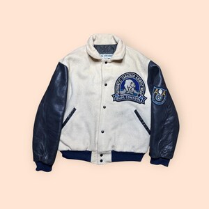 May include: Cream-colored varsity jacket with navy blue leather sleeves. Features a circular patch with a polar bear and the text "Authentic Canadian Arctic Wear Dual Control". Includes a navy blue collar, cuffs, and snap closures.