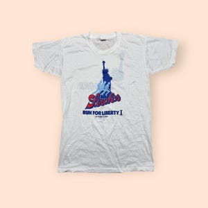 May include: White t-shirt featuring a graphic of the Statue of Liberty in blue, with the text "Stroh's" in red and blue, and "RUN FOR LIBERTY I OCTOBER 13 1984" in blue. The shirt has a crew neck and short sleeves.