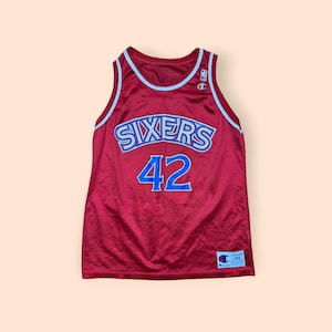 May include: A maroon basketball jersey with the word "SIXERS" in light blue and silver lettering across the chest, and the number "42" in blue. The jersey has light blue trim around the neck and armholes, and a Champion logo.