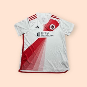 May include: White soccer jersey with red accents and the "United Healthcare" logo. The jersey features the Adidas logo and a team emblem on the chest. Red stripes run down the sides and across the shoulders.