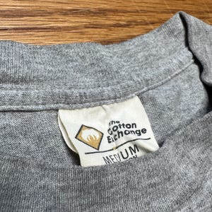May include: Close-up of a gray t-shirt collar with a tag. The tag reads "the Cotton Exchange" and "MEDIUM". The tag has a diamond-shaped logo with a design inside. The shirt is made of a soft, knit fabric.