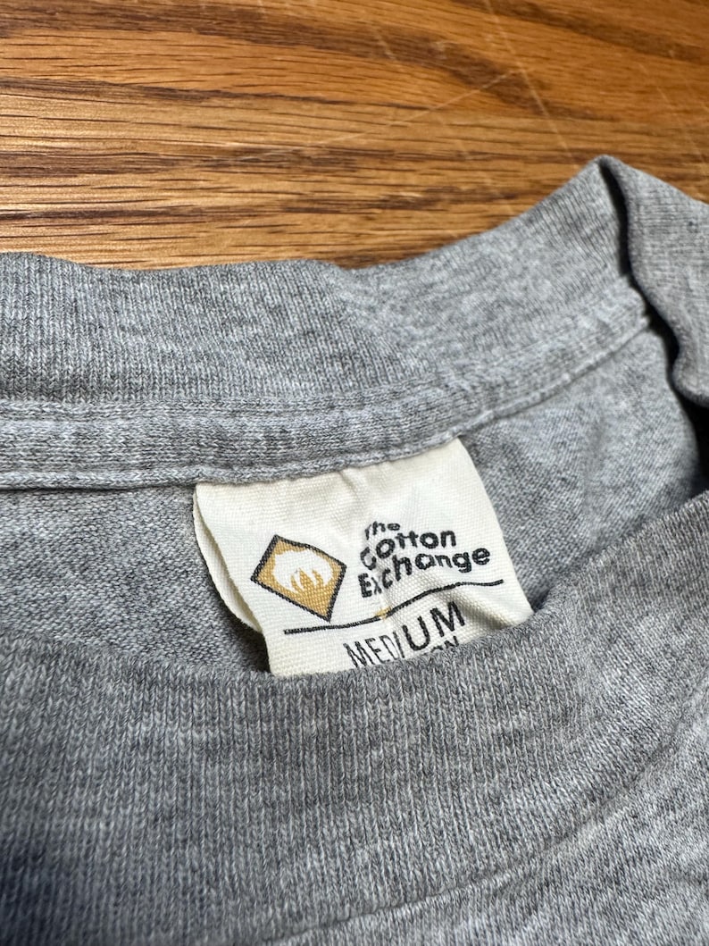 May include: Close-up of a gray t-shirt collar with a tag. The tag reads "the Cotton Exchange" and "MEDIUM". The tag has a diamond-shaped logo with a design inside. The shirt is made of a soft, knit fabric.