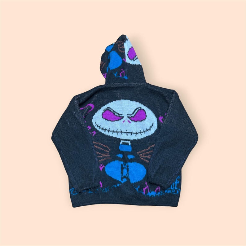 Puede incluir: Una sudadera de punto negra con capucha que presenta un gran gr&aacute;fico central de un personaje de calavera blanca con ojos morados y boca cosida. El dise&ntilde;o incluye detalles en azul y morado, con im&aacute;genes adicionales en la capucha y las mangas.