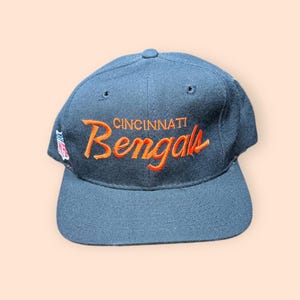 May include: A navy blue Cincinnati Bengals baseball cap. The team name "Bengals" is embroidered in orange script across the front, with "Cincinnati" above it. The cap has a curved brim and a button on top.