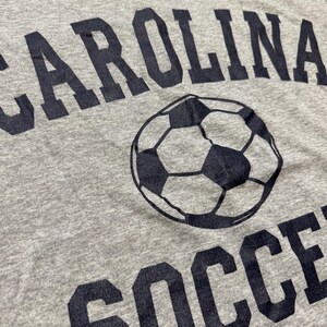 May include: Gray t-shirt with the words "CAROLINA SOCCER" printed in dark blue. A soccer ball graphic is centered between the words. The shirt has a crew neck and is made of a soft, textured fabric.