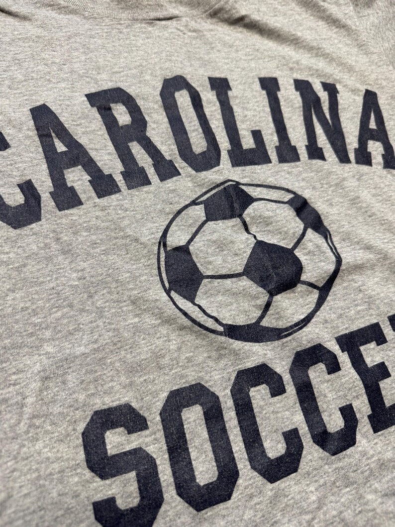 May include: Gray t-shirt with the words "CAROLINA SOCCER" printed in dark blue. A soccer ball graphic is centered between the words. The shirt has a crew neck and is made of a soft, textured fabric.