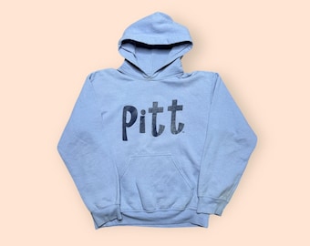 May include: A light blue hooded sweatshirt with the word "Pitt" printed in dark blue across the chest. The sweatshirt has a front pocket and a drawstring hood. The background is a light peach color.