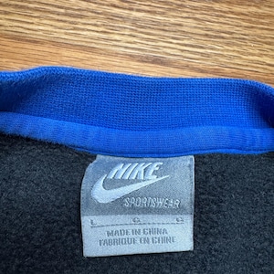 Puede incluir: Primer plano de una sudadera negra con cuello azul. El logotipo de Nike y "Sportswear" est&aacute;n impresos en una etiqueta blanca. La etiqueta tambi&eacute;n dice "Made in China / Fabriqu&eacute; en Chine".