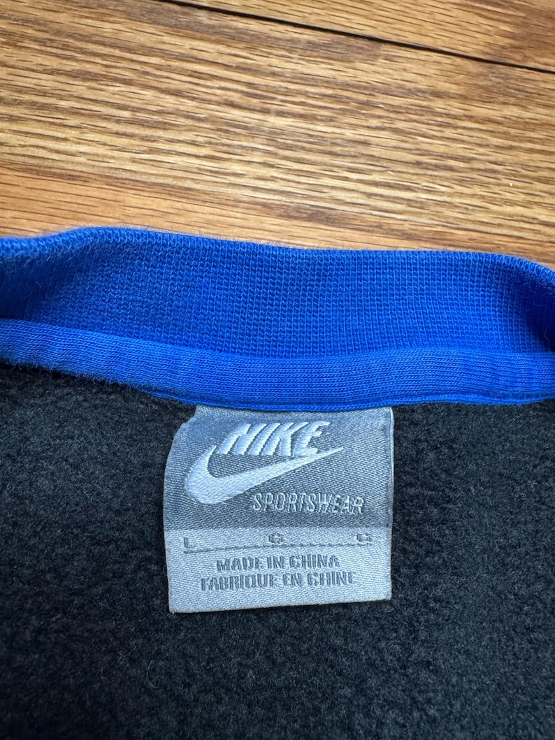 Puede incluir: Primer plano de una sudadera negra con cuello azul. El logotipo de Nike y "Sportswear" est&aacute;n impresos en una etiqueta blanca. La etiqueta tambi&eacute;n dice "Made in China / Fabriqu&eacute; en Chine".
