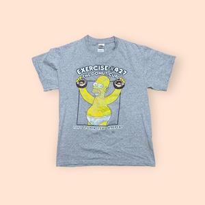 May include: A gray t-shirt featuring a cartoon image of Homer Simpson lifting donuts as weights. The text on the shirt reads "Exercise #427 The Donut Curl 1. Lift 2. Open 3. Eat 4. Repeat."