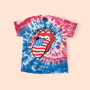 May include: A short-sleeve t-shirt with a red, white, and blue tie-dye pattern. The center features The Rolling Stones tongue and lips logo, with the tongue filled in with an American flag design and the text The Rolling Stones arched above.