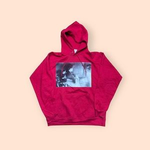 May include: A red hooded sweatshirt with a front pocket and a black and white image on the front. The image appears to be a photograph of people. The sweatshirt is made of a soft material.