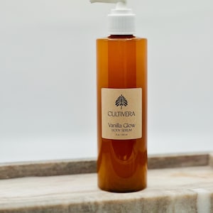 Op de afbeelding: Een bruine body serumfles met een witte pompdosering. Het etiket luidt "Cultivera Vanilla Glow Body Serum" en is 240 ml. De fles staat op een marmeren dienblad.