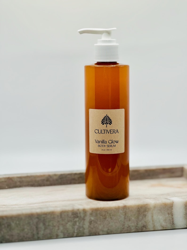 Op de afbeelding: Een bruine body serumfles met een witte pompdosering. Het etiket luidt "Cultivera Vanilla Glow Body Serum" en is 240 ml. De fles staat op een marmeren dienblad.