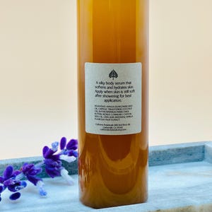 Op de afbeelding: Een heldere fles lichaamsserum met een lichtblauwe dop. De amberkleurige vloeistof is gelabeld met tekst die de voordelen en ingredi&euml;nten van het product beschrijft. De fles staat op een grijze tray met paarse bloemen.