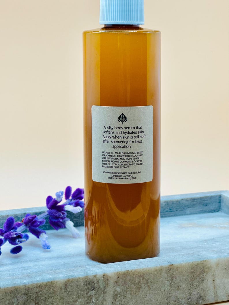Op de afbeelding: Een heldere fles lichaamsserum met een lichtblauwe dop. De amberkleurige vloeistof is gelabeld met tekst die de voordelen en ingredi&euml;nten van het product beschrijft. De fles staat op een grijze tray met paarse bloemen.