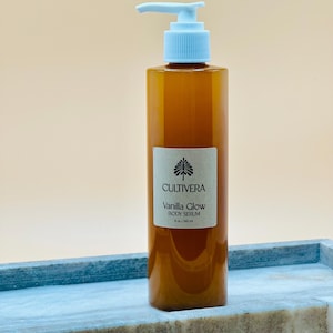 Op de afbeelding: Een bruine body serumfles met een lichtblauwe pomp, met het label "Cultivera Vanilla Glow Body Serum". De fles is 240 ml. De fles staat op een blauw en grijs marmeren dienblad.