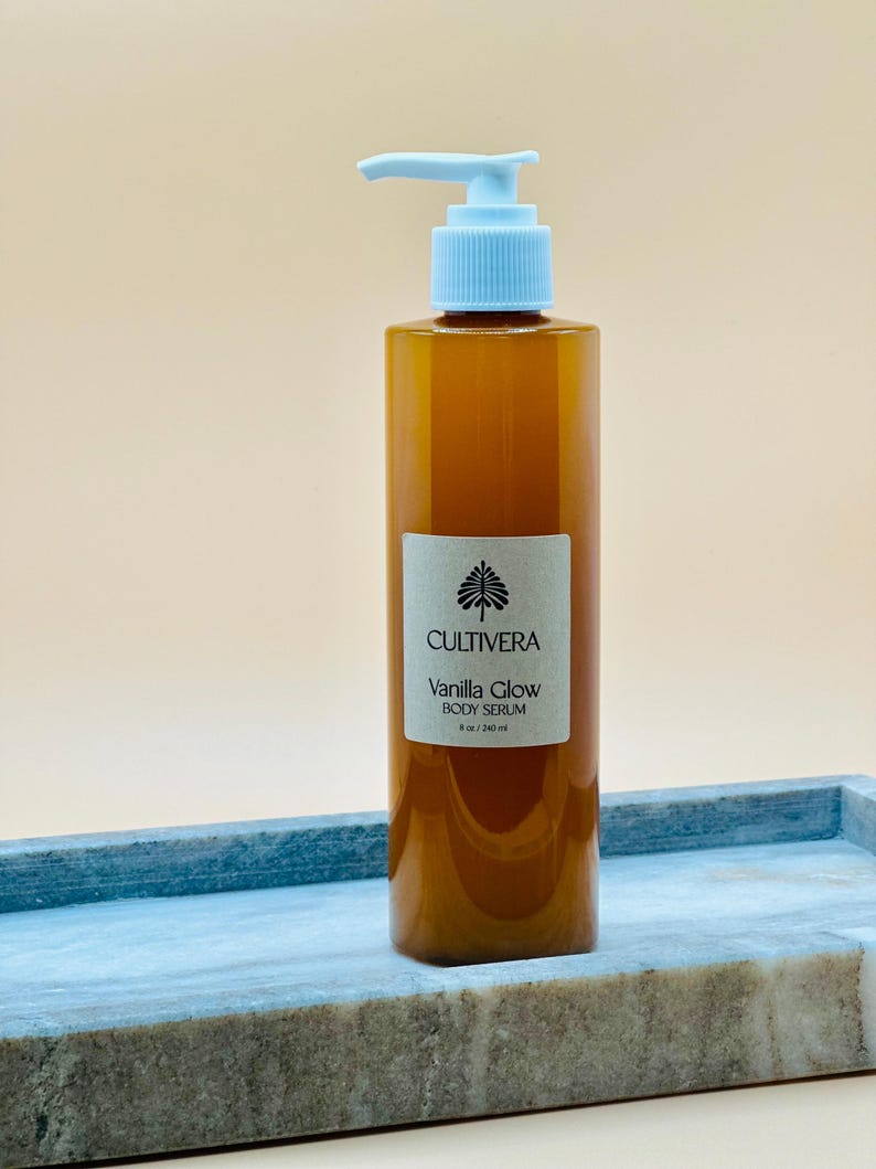 Op de afbeelding: Een bruine body serumfles met een lichtblauwe pomp, met het label "Cultivera Vanilla Glow Body Serum". De fles is 240 ml. De fles staat op een blauw en grijs marmeren dienblad.