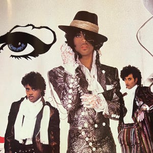 May include: Image of Prince and band members. Prince is wearing a black hat with a gold band, a patterned jacket, and lace gloves. The image has a white background with a large eye graphic. Other band members are also wearing patterned jackets.