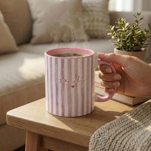 May include: A pink and white striped coffee mug filled with coffee, with steam rising from the top. The mug has the name "Sandy" written on it in cursive with a small heart. The mug is held by a person.