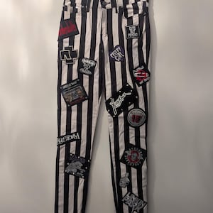 Può includere: Jeans a righe bianche e nere con diverse toppe con loghi di band e frasi come "Cute But Psycho" e "Creepy Girl". I jeans sono appesi a una gruccia contro un muro bianco.