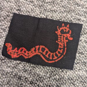 Può includere: Una toppa in tessuto nero con un disegno di serpente rosso stilizzato. Il serpente ha un corpo segmentato e una testa dettagliata. La toppa è rettangolare e sembra essere fatta di un materiale tessuto.