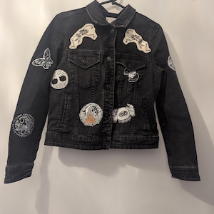Peut inclure: Veste en jean noire ornée de divers écussons brodés. Les écussons comprennent un papillon, un arbre de vie, un crâne et d'autres motifs gothiques. La veste est suspendue à un cintre noir contre un mur blanc.