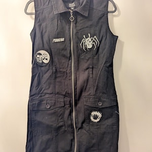 Könnte beinhalten: Schwarzes ärmelloses Jeanskleid mit Reißverschluss und mehreren Taschen. Mit Aufnähern mit Schädel- und Spinnenmotiven und dem Wort "PUNKERS". Das Kleid hängt an einem schwarzen Kleiderbügel.