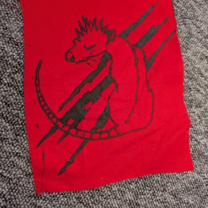 Op de afbeelding: Een rode stoffen patch met een zwarte illustratie van een rat met een stekelige kroon en een gebogen staart. Drie zwarte klauwstrepen staan naast de rat. De patch is rechthoekig met zichtbare stiksels.