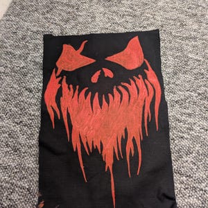 Puede incluir: Tela negra con un diseño de rostro estilizado en rojo. El rostro tiene ojos grandes, una nariz y una boca dentada en forma de llama. La obra de arte está centrada en la tela, creando un contraste visual llamativo.