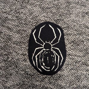 May include: Black oval patch featuring a white spider design. The spider has eight legs and a rounded body. The patch is made of a dark fabric and is likely an iron-on or sew-on patch for clothing or accessories.