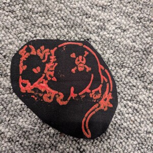 May include: Black patch with a red design featuring a skull and crossbones and other stylized elements. The patch has an irregular shape and is likely made of fabric.