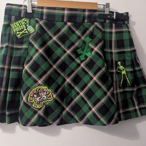 May include: A green and black plaid pleated skirt featuring a side buckle strap and several embroidered patches, including a bottle labelled DEATH POISON, a green cross, a skeleton figure, and a kitten with the text anarchist.