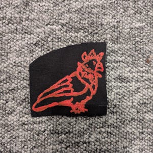 May include: A black fabric patch featuring a red embroidered bird design. The bird is stylized with a crown and a long tail. The patch is rectangular with a curved edge.