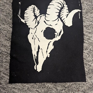 May include: Black rectangular patch with a white ram skull design. The skull has large, curved horns and detailed bone structure. The patch is likely made of fabric and is suitable for sewing or ironing onto clothing or accessories.